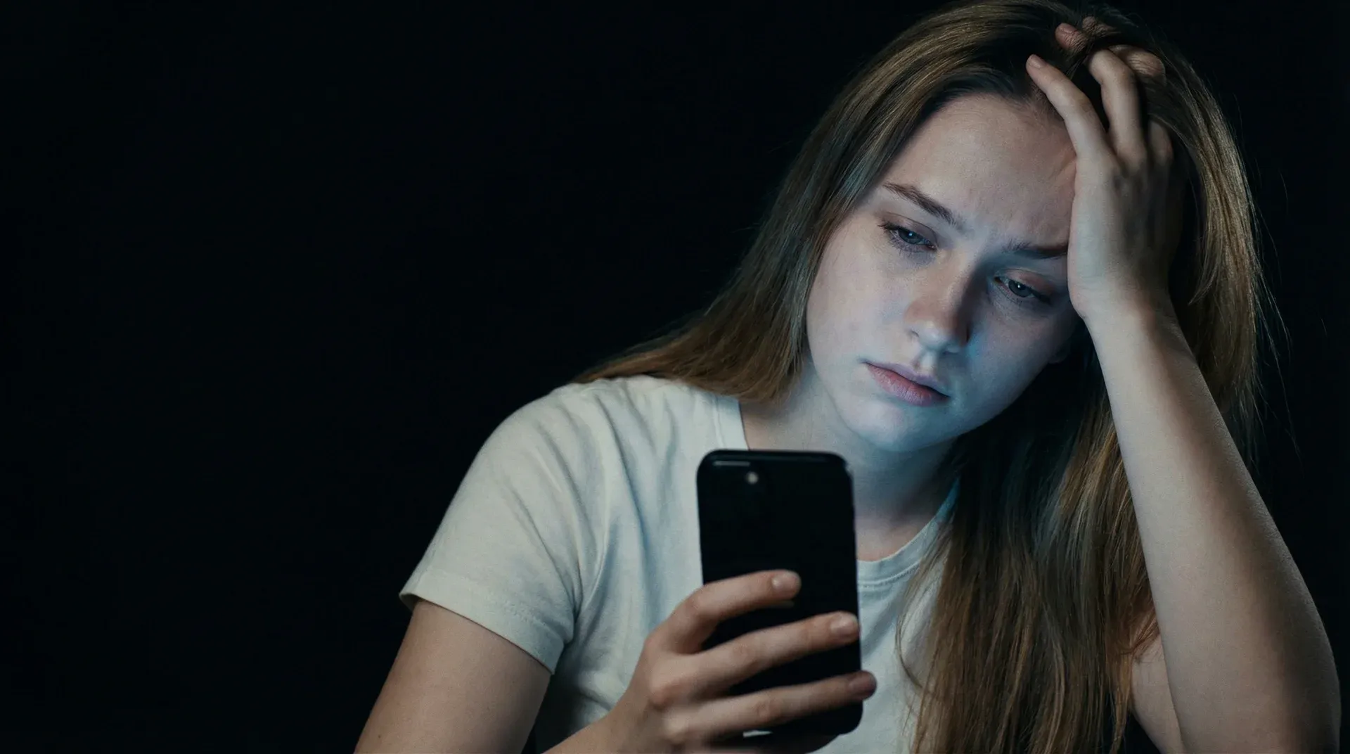Person sitting alone in a dark room holding their phone with a frustrated expression
