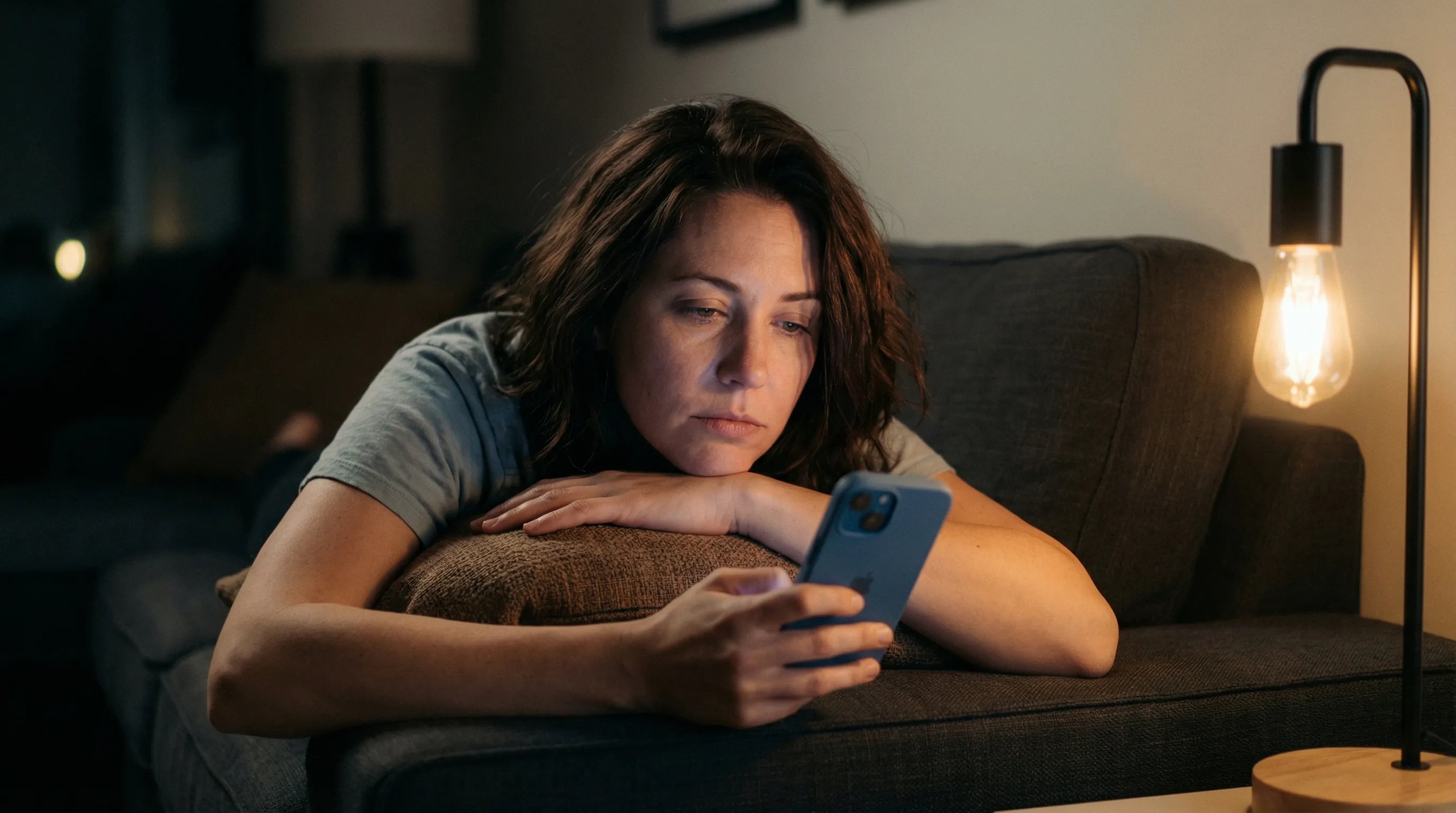 Woman lying on couch at night scrolling through smartphone in dim lighting